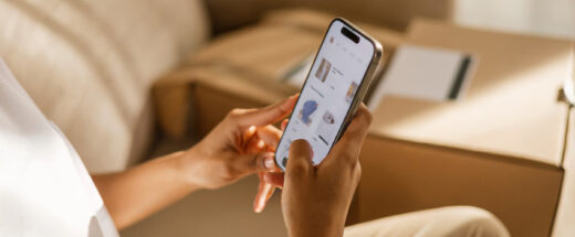 Person holding a smartphone and browsing an online store with clothing items displayed, with cardboard boxes in the background