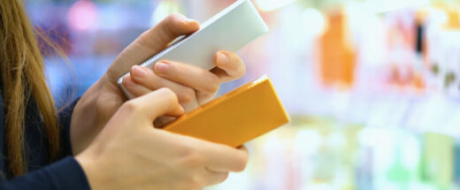 A person's hands holding two smartphones - one white and one orange/yellow - against a blurred colorful background, suggesting comparison shopping or mobile device selection.