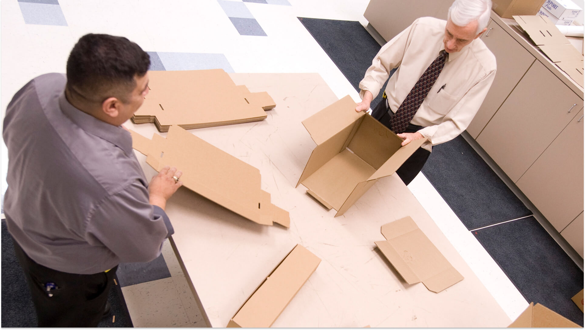 Two business professionals examining flat-pack cardboard box prototypes on office floor for packaging design review