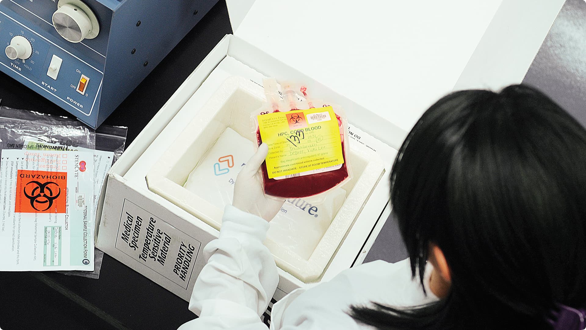 A laboratory technician in a white coat examines a blood sample container with biohazard labeling inside a white styrofoam shipping box.