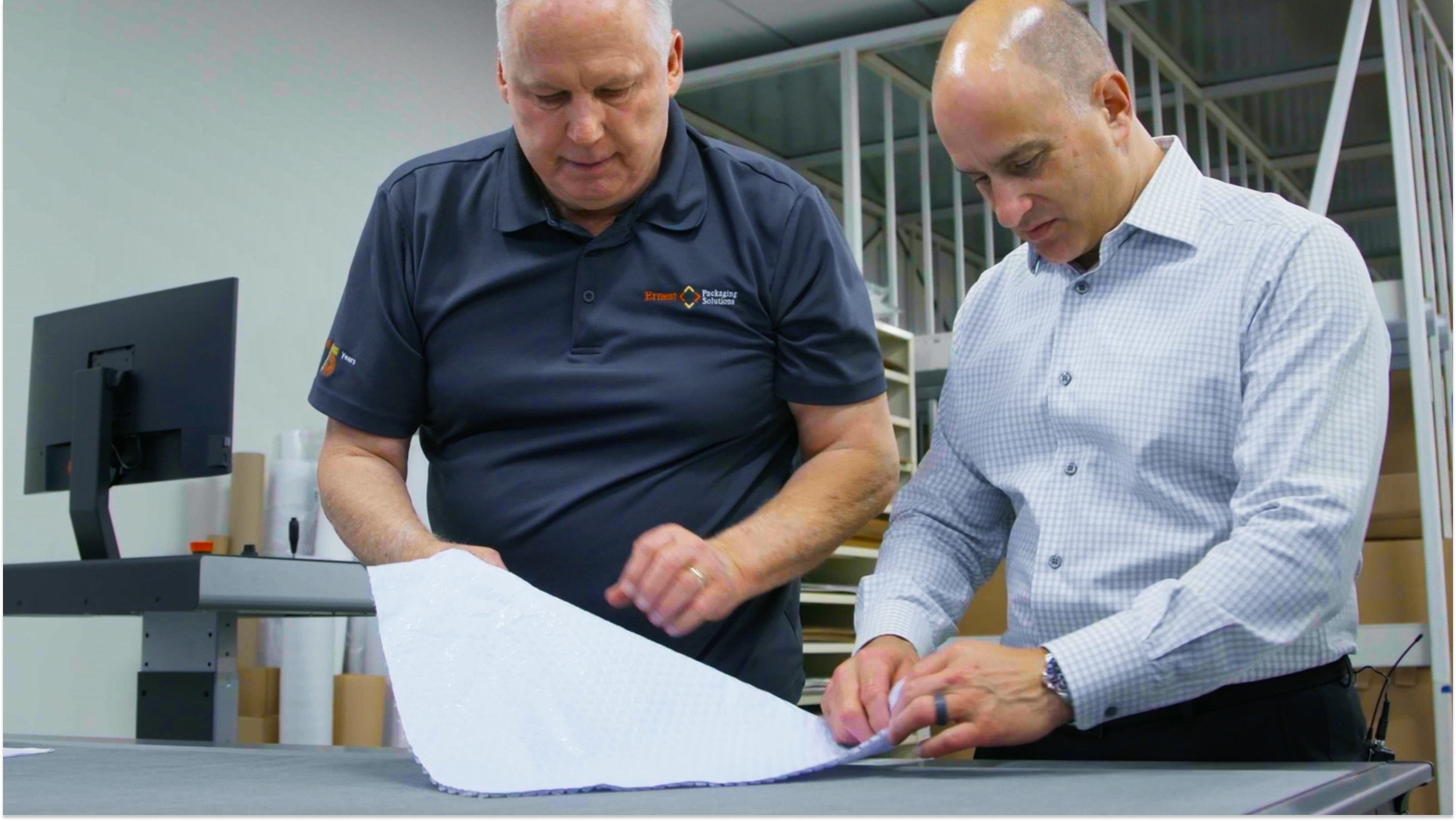 Two packaging professionals examining white paperboard material sample on table in modern packaging facility